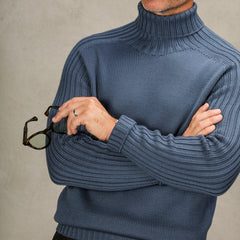 A person in an Airforce Blue Rain Wool Rollneck from Gran Sasso crosses their arms and holds a pair of glasses.