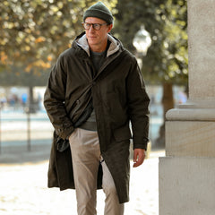 An individual dressed in an Aspesi Military Green Cotton Nylon Thermore Rams Parka, along with a gray hoodie, beanie, and glasses, is walking outdoors near a tree and a column.