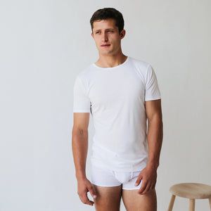 A man wearing an Earth Organic Pima Cotton Crewneck T-shirt from The White Briefs and matching white underwear stands beside a wooden stool against a plain background.