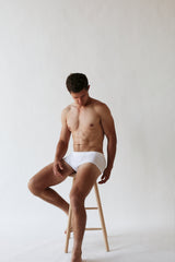 A man wearing The White Briefs Elm Organic Pima Cotton Mid Briefs, in a regular fit and white color, sits on a wooden stool against a plain background, gazing downwards.