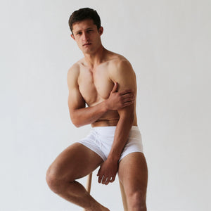 A person wearing Wil Organic Pima Cotton Trunks from The White Briefs sits on a stool against a plain background, with one hand on their opposite shoulder.