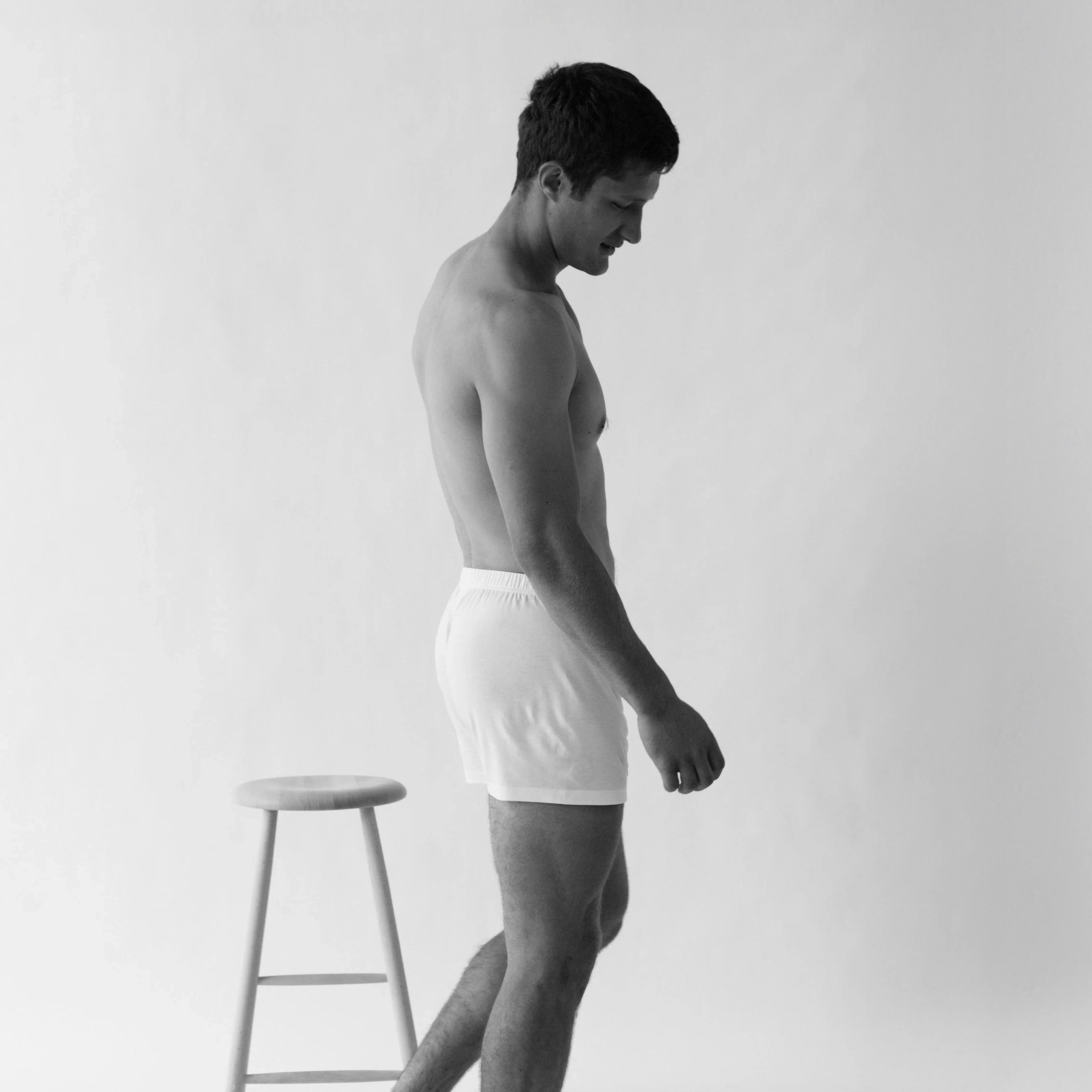 A man in profile wearing Henner Cotton Lyocell Boxer Shorts by The White Briefs stands beside a wooden stool against a plain background.