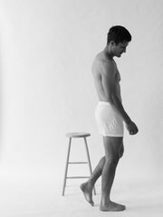 A man wearing The White Briefs' Henner Cotton Lyocell Boxer Shorts, featuring an elastic waistband, stands beside a stool against a plain backdrop.