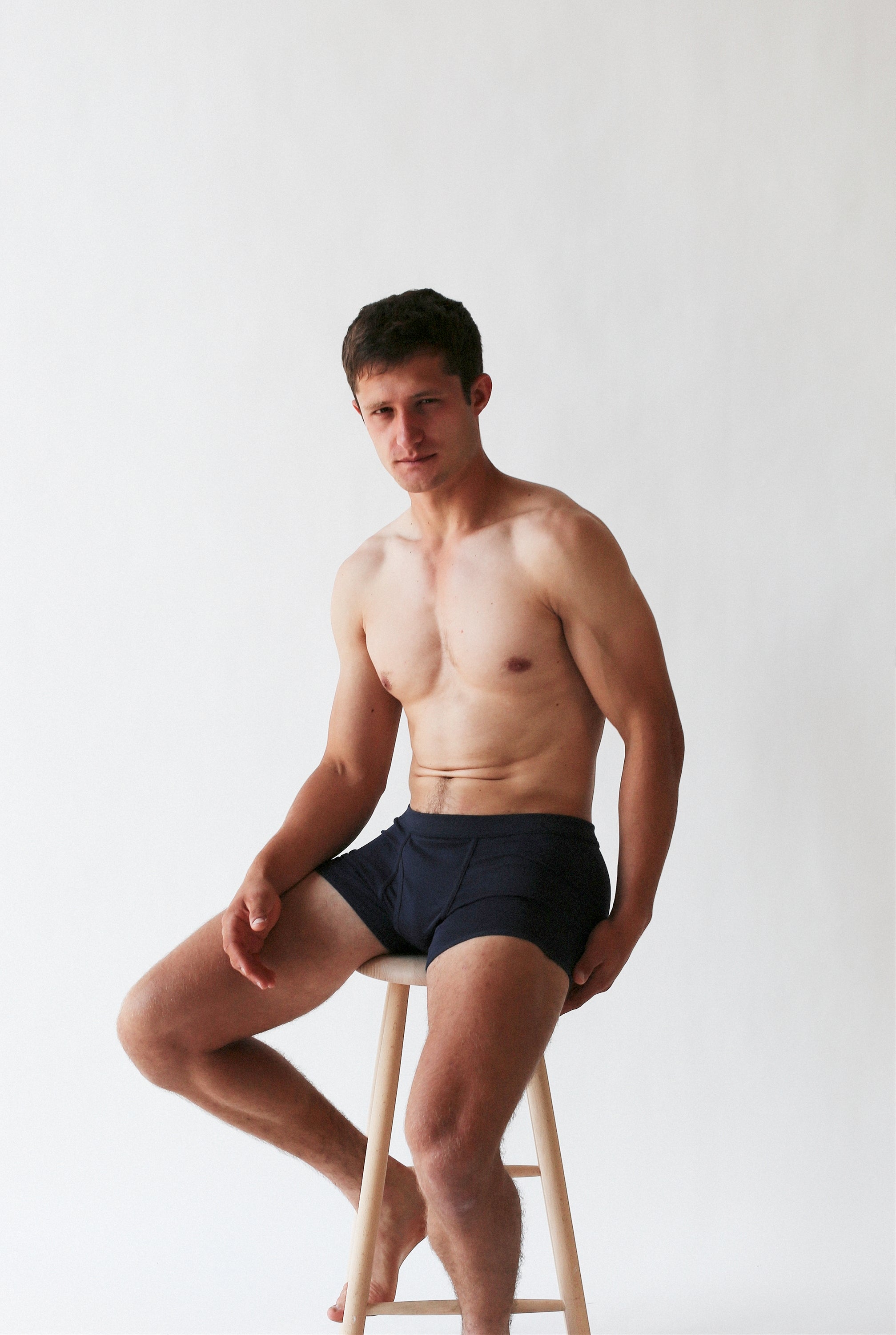 A person wearing Wil Organic Pima Cotton Trunks by The White Briefs, in classic black, sits on a wooden stool against a plain white background.