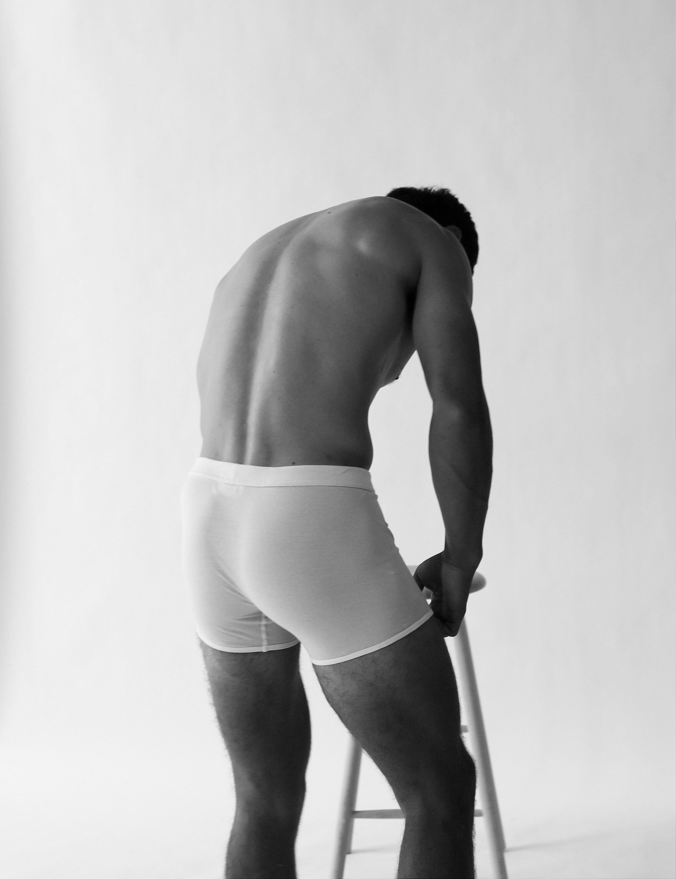 A person wearing The White Briefs' Wil Organic Pima Cotton Trunks in regular fit, white, stands beside a wooden ladder, facing away against a plain background.