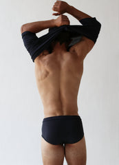 A person facing away, taking off a dark shirt, is wearing The White Briefs' Platan Organic Pima Cotton Classic Briefs against a neutral background.