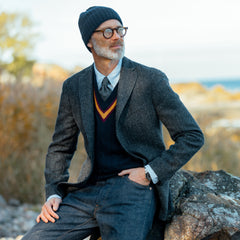 A man with a beard, wearing a beanie and glasses, sits on a rock outdoors while sporting a Boglioli Dark Grey Herringbone Wool Tweed K Jacket made with unstructured craftsmanship over his sweater.