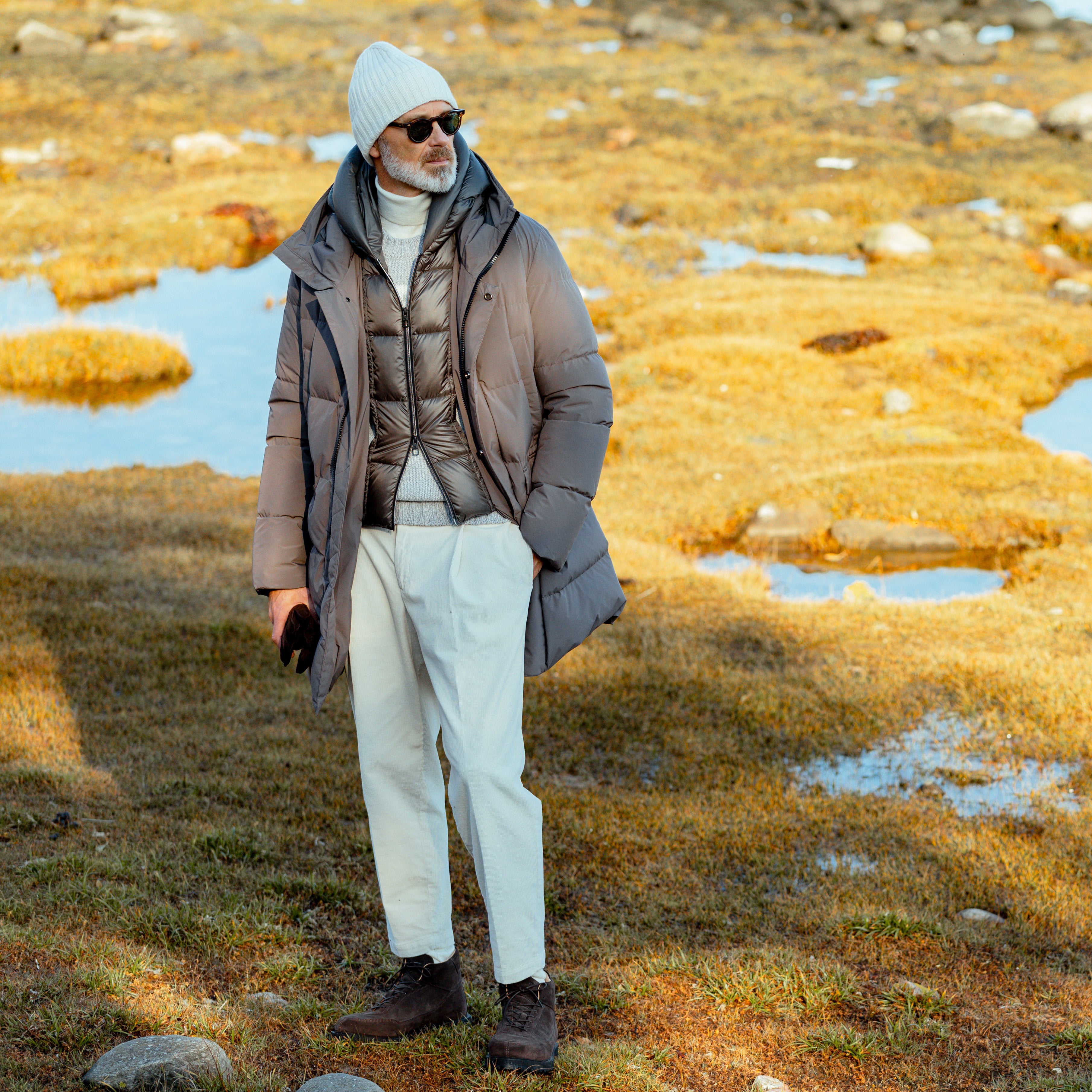 A person in a gray winter coat and white beanie stands on grassy terrain with scattered puddles, under a clear sky, comfortably wearing regular fit Off White Corduroy Courmayeur Trousers by Briglia.