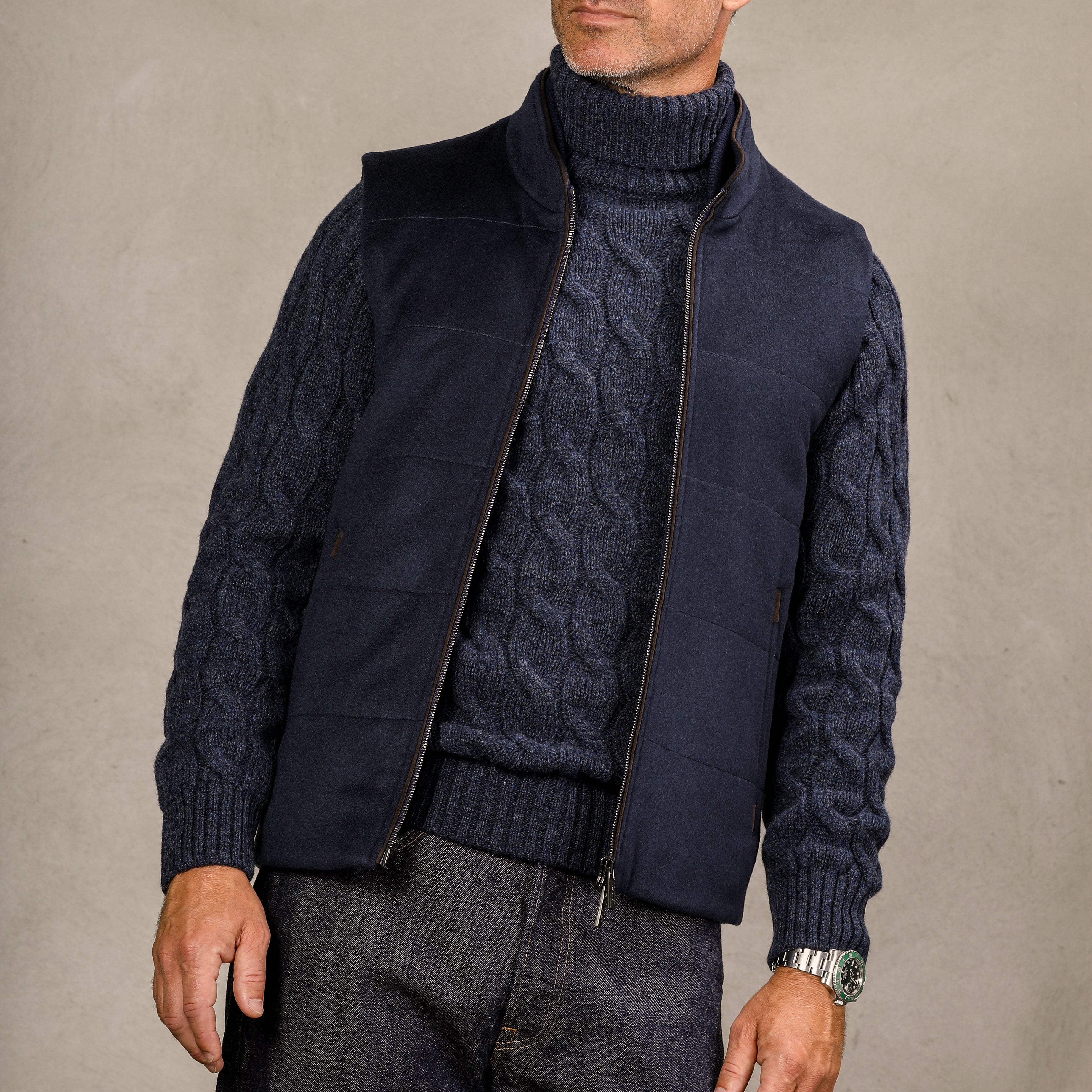 A person wearing a Gran Sasso Dark Blue Wool Cable Knit Rollneck, a navy vest, and dark jeans, with their left hand resting by their side, against a plain background.