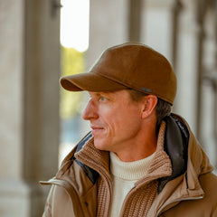 A person wearing a brown jacket featuring a Loro Piana Storm System, a Camel Loro Piana Wool Cashmere Baseball Cap by Wigéns, and a knitted sweater stands in an outdoor setting, looking to the side.