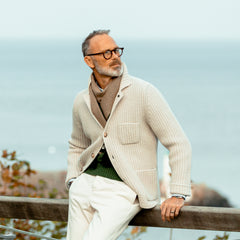 A man wearing a Gran Sasso Cream Beige Chunky Knitted Wool Cardigan and glasses stands by a wooden railing, gazing into the distance with the sea in the background.