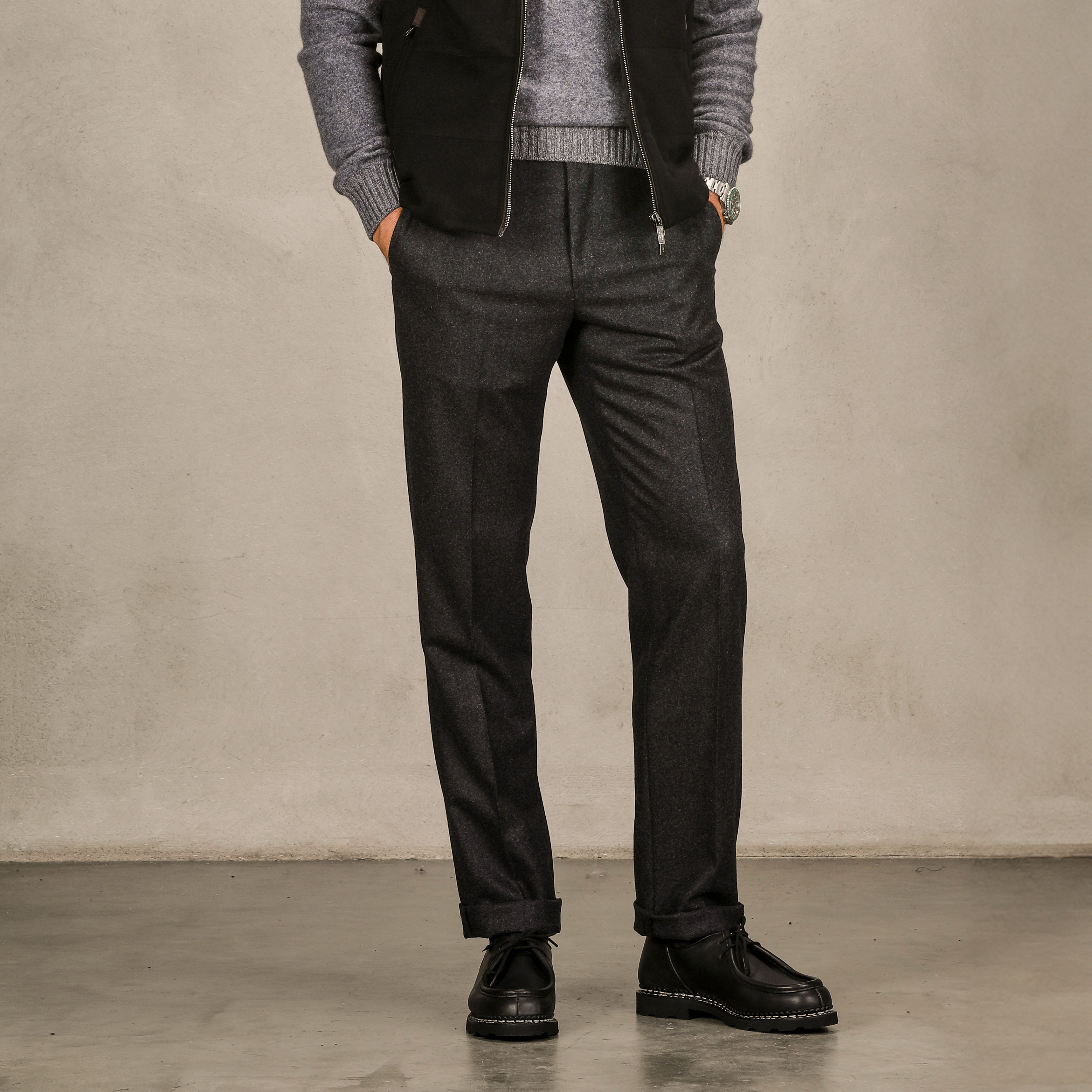 A person stands against a plain background, wearing a gray sweater and Luigi Bianchi Charcoal Grey Wool Flannel Flat Front Trousers. Only the lower half of the body is visible. Hands are in pockets, completing the look with dark pants and black shoes.