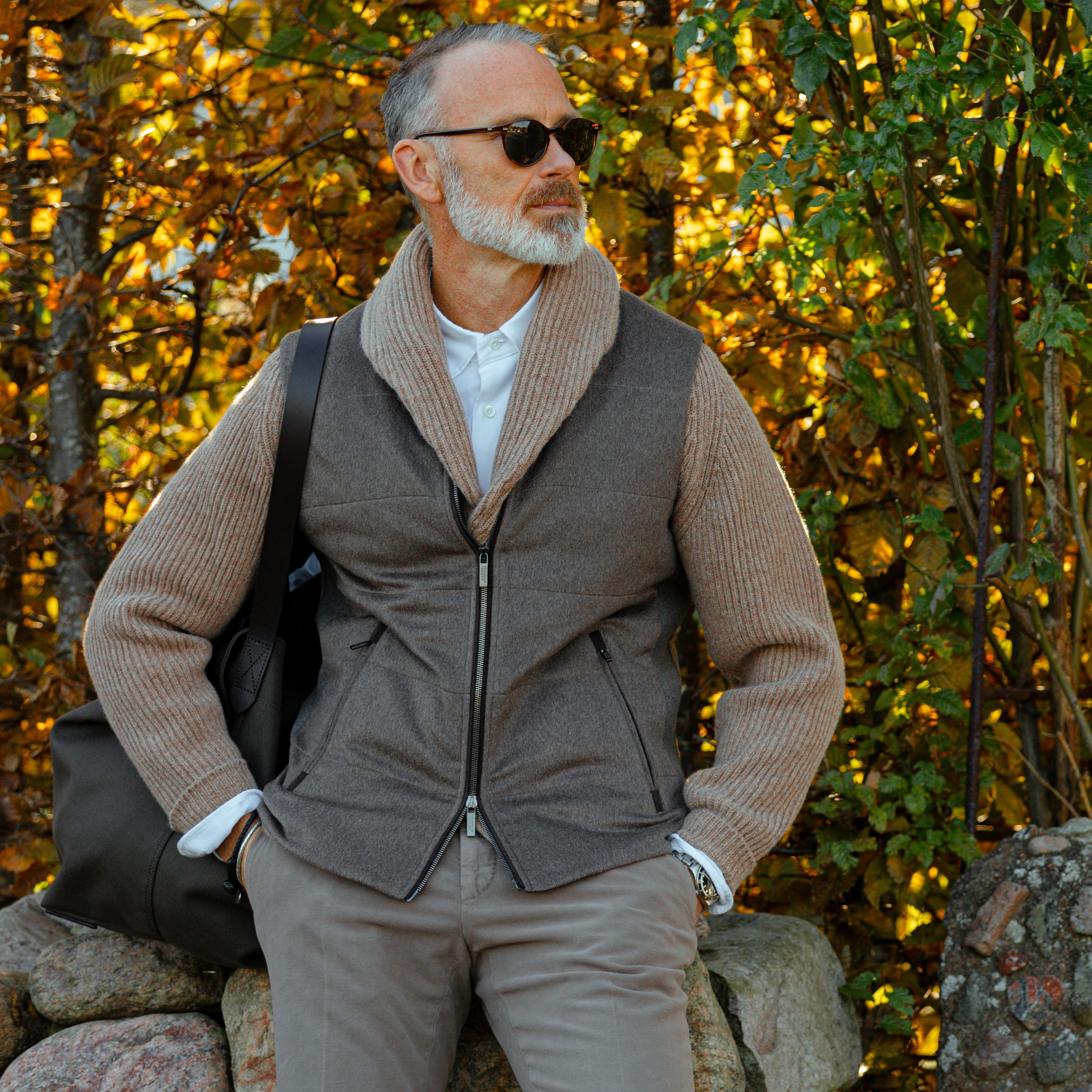 A man in sunglasses sits on a stone wall, wearing a Colt Beige Lambswool Shawl Collar Cardigan by William Lockie with leather buttons over a white shirt and gray pants. He's holding a dark bag, with autumn foliage in the background.