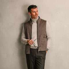 A man leans against a plain wall, wearing a light turtleneck, a Dark Taupe Water Repellent Cashmere Gilet by Maurizio Baldassari, and holding eyeglasses in one hand.