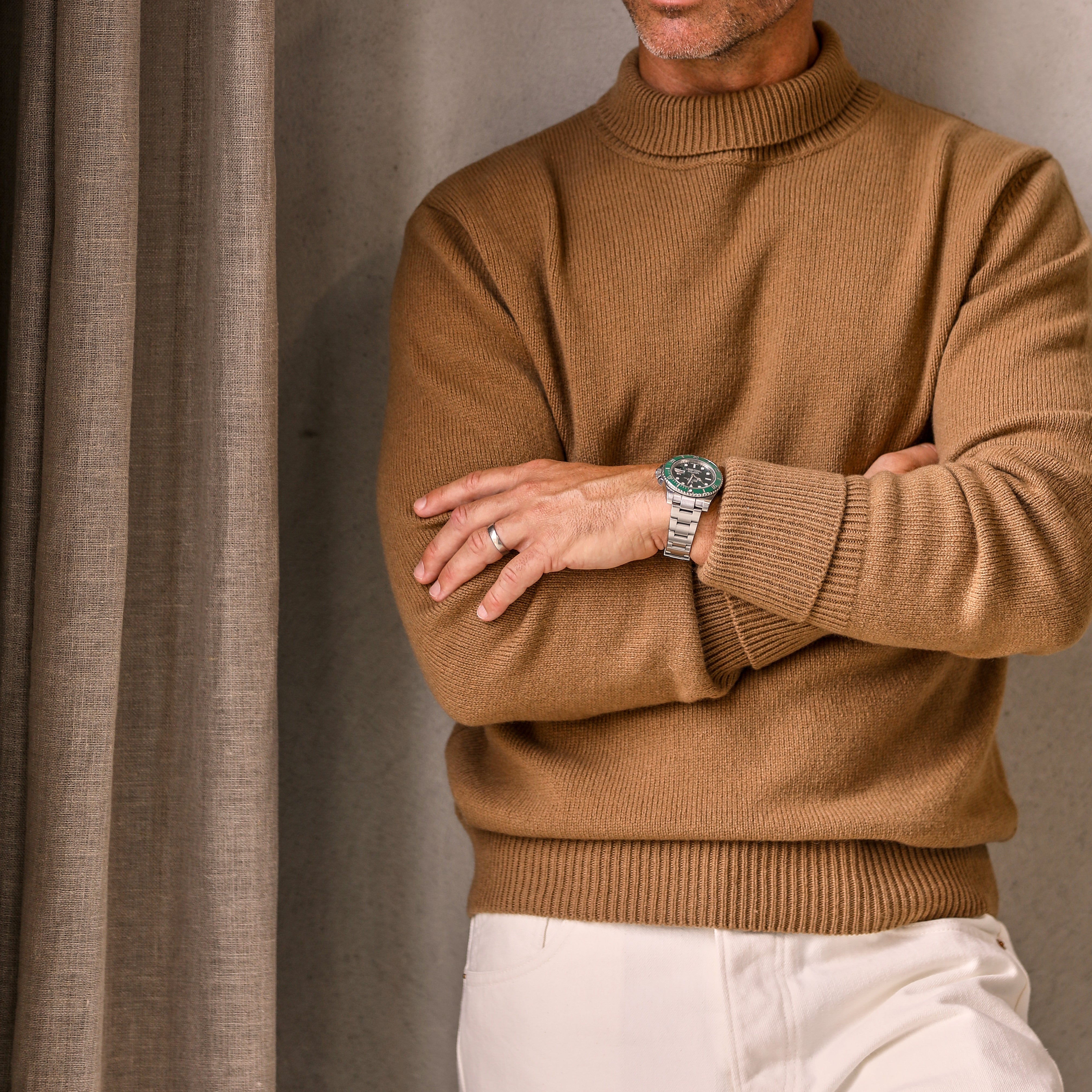 A man wearing a G.R.P Camel Beige Wool Cashmere Mock Neck Sweater and white pants stands with arms crossed, displaying a wristwatch and a wedding ring.