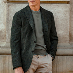 A person in a Green Herringbone Wool Tweed K Jacket by Boglioli, crafted with unstructured flair, layered over a gray sweater, stands against a stone wall. Their face is not visible.