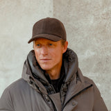 A man wearing a Fedeli Dark Brown Felted Cashmere Cap and a gray puffer jacket stands against a textured wall, looking into the distance.