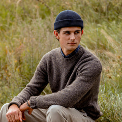 A person wearing a brown merino wool sweater from Nitto, a Marine Blue Merino Wool Ara Beanie, and beige pants sits outdoors amidst tall grass, looking to the side.