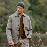 A person wearing a Khaki Green Merino Wool Ara Beanie from Nitto, along with a grey jacket and brown sweater, walks outside in a natural setting.