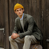 A person wearing an Ocre Yellow Merino Wool Ara Beanie from Nitto, a grey blazer, and beige trousers sits on a stone in front of a wooden wall, looking to the left.