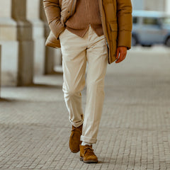 A person walks along a paved path, dressed in a brown jacket, brown sweater, and Luigi Bianchi Light Beige Cotton Twill Flat Front Trousers paired with matching brown shoes.