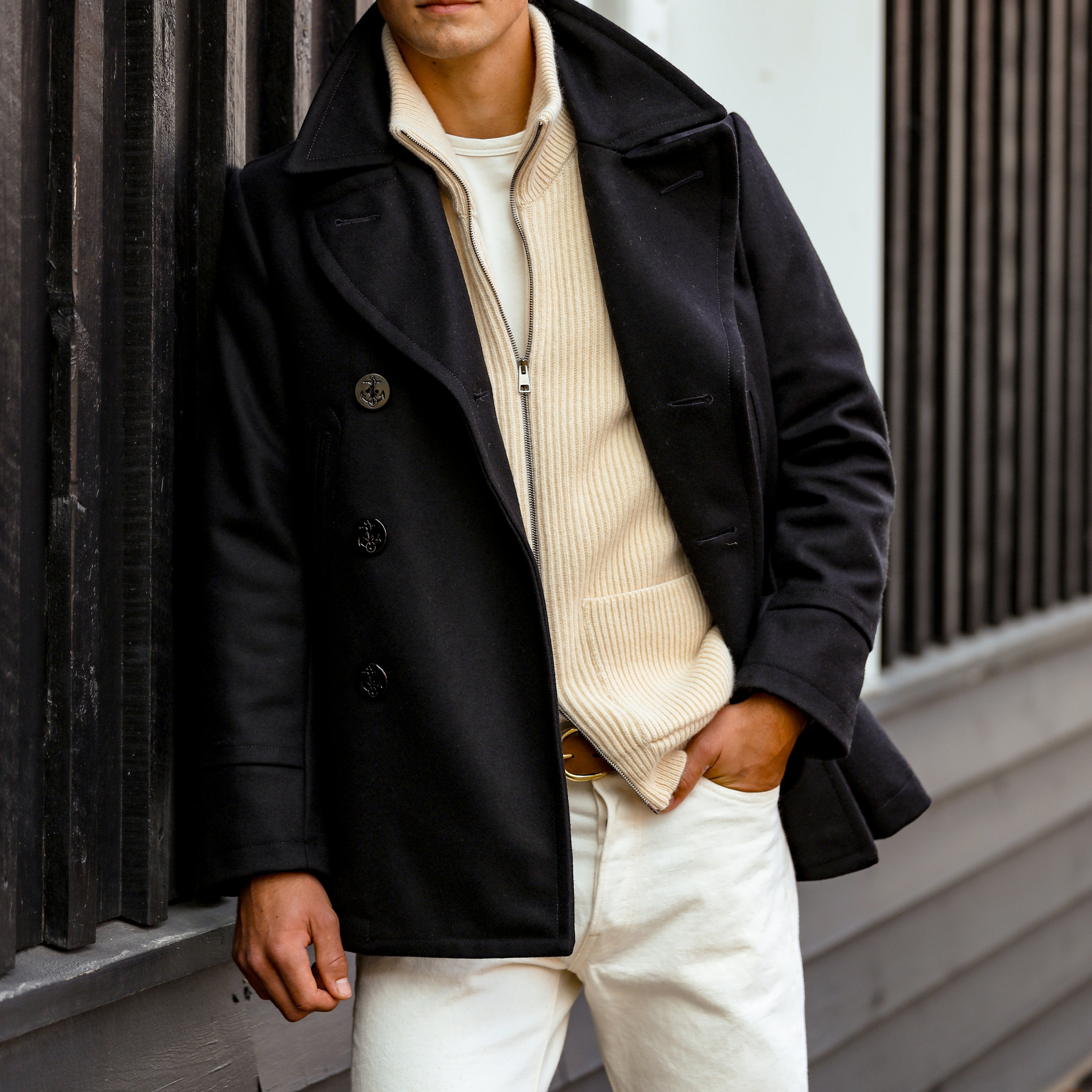 A man wearing a Dark Navy Heavy Wool Peacoat over a beige sweater and white pants, crafted by Manifattura Ceccarelli, stands in front of a dark vertical slat wall with one hand in his pocket.