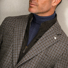 A man with facial hair is wearing a houndstooth blazer, a black zip-up jacket, and a Petrol Blue Merino Wool Roll Neck sweater by Gran Sasso. The background features a neutral-colored wall.