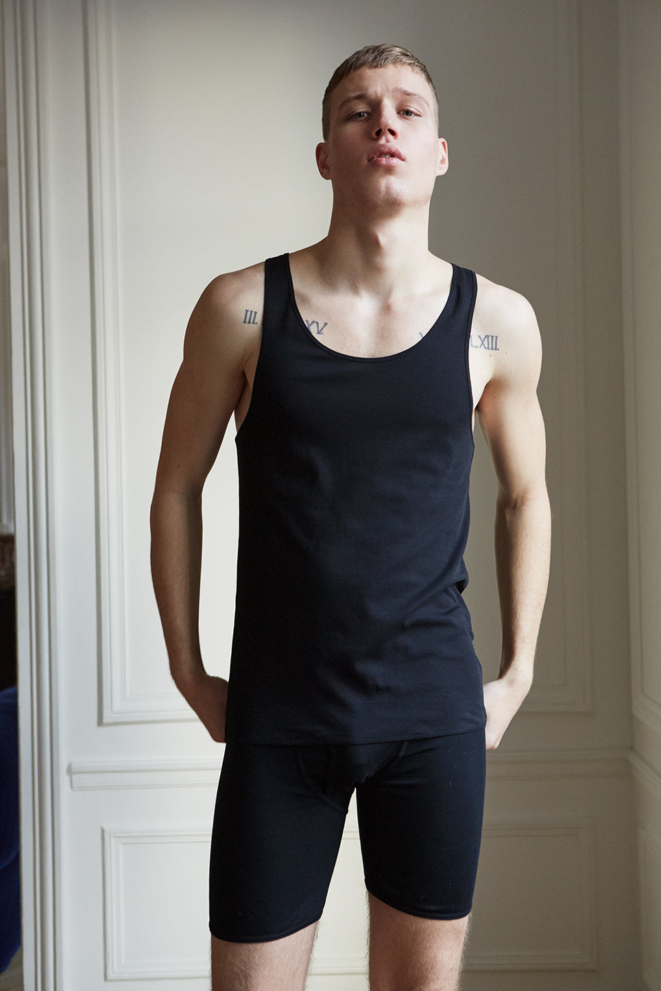 An individual wearing a Rye Organic Pima Cotton Singlet by The White Briefs and shorts, both crafted from eco-friendly fabric, standing in front of a white-paneled wall.