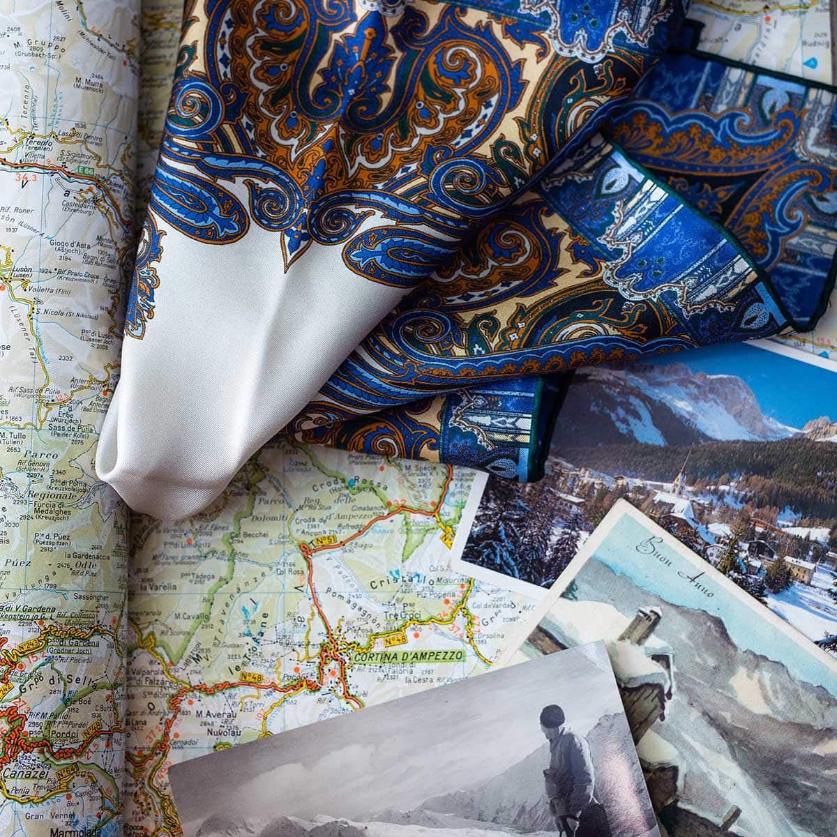 The Cortina Blue Silk Pocket Square by Sera Fine Silk, featuring detailed blue and gold patterns and crafted from pure Como silk, is draped over a map. Two black-and-white photos of a snowy landscape and people in winter attire are placed on the map. The luxurious pocket square, with its hand-rolled edges, adds an elegant touch to the scene.