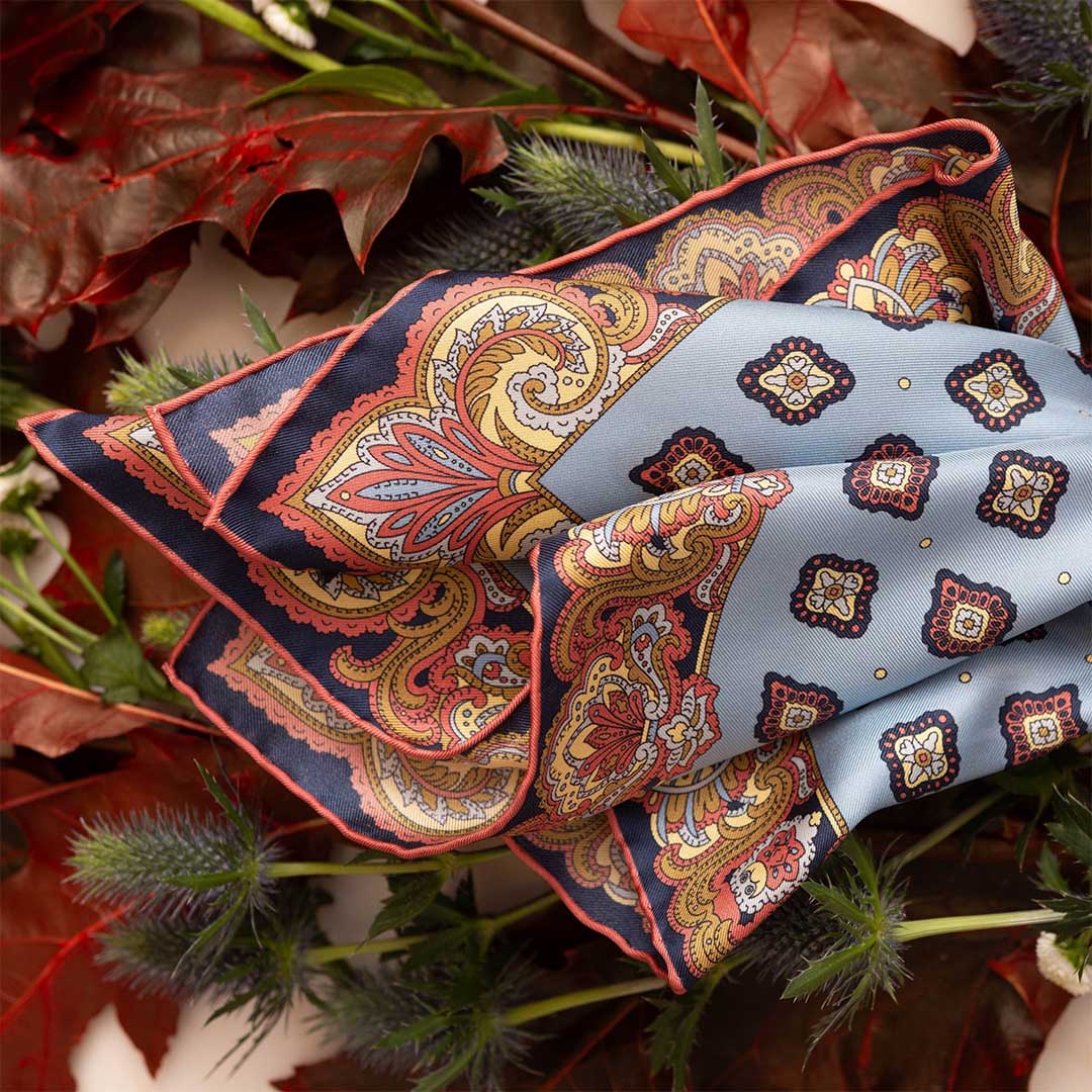 A Cardo Light Blue Silk Pocket Square by Sera Fine Silk, boasting a paisley pattern in blue and orange, is laid out over a bed of assorted leaves and flowers.