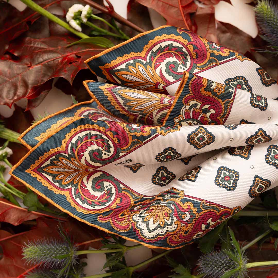 The Bucaneve White Silk Pocket Square by Sera Fine Silk is a square scarf showcasing intricate patterns with vibrant red, green, and yellow hues. It is crafted from pure Como silk and adorned with autumn leaves and Snowdrop flowers in the background.