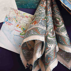 The Vernazza Cream Silk Pocket Square from Sera Fine Silk, crafted from pure Como silk with hand-rolled edges, is placed on a table with a colorful map beneath it.