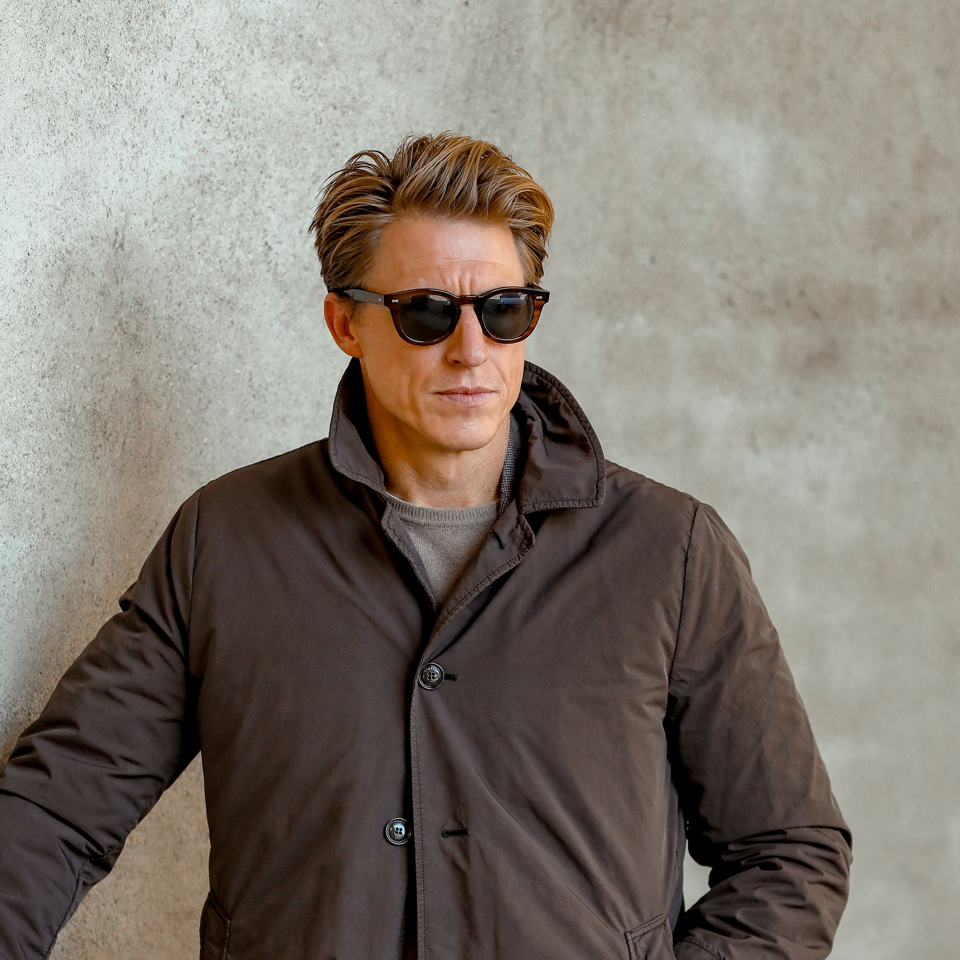 A man with short brown hair wearing The Bespoke Dudes Donegal Earth sunglasses with Bio Gradient Grey Lenses (49mm) and a brown coat stands against a textured concrete wall, with his hands in his pockets.