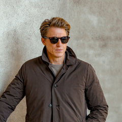 A man with short brown hair wearing The Bespoke Dudes Donegal Earth sunglasses with Bio Gradient Grey Lenses (49mm) and a brown coat stands against a textured concrete wall, with his hands in his pockets.