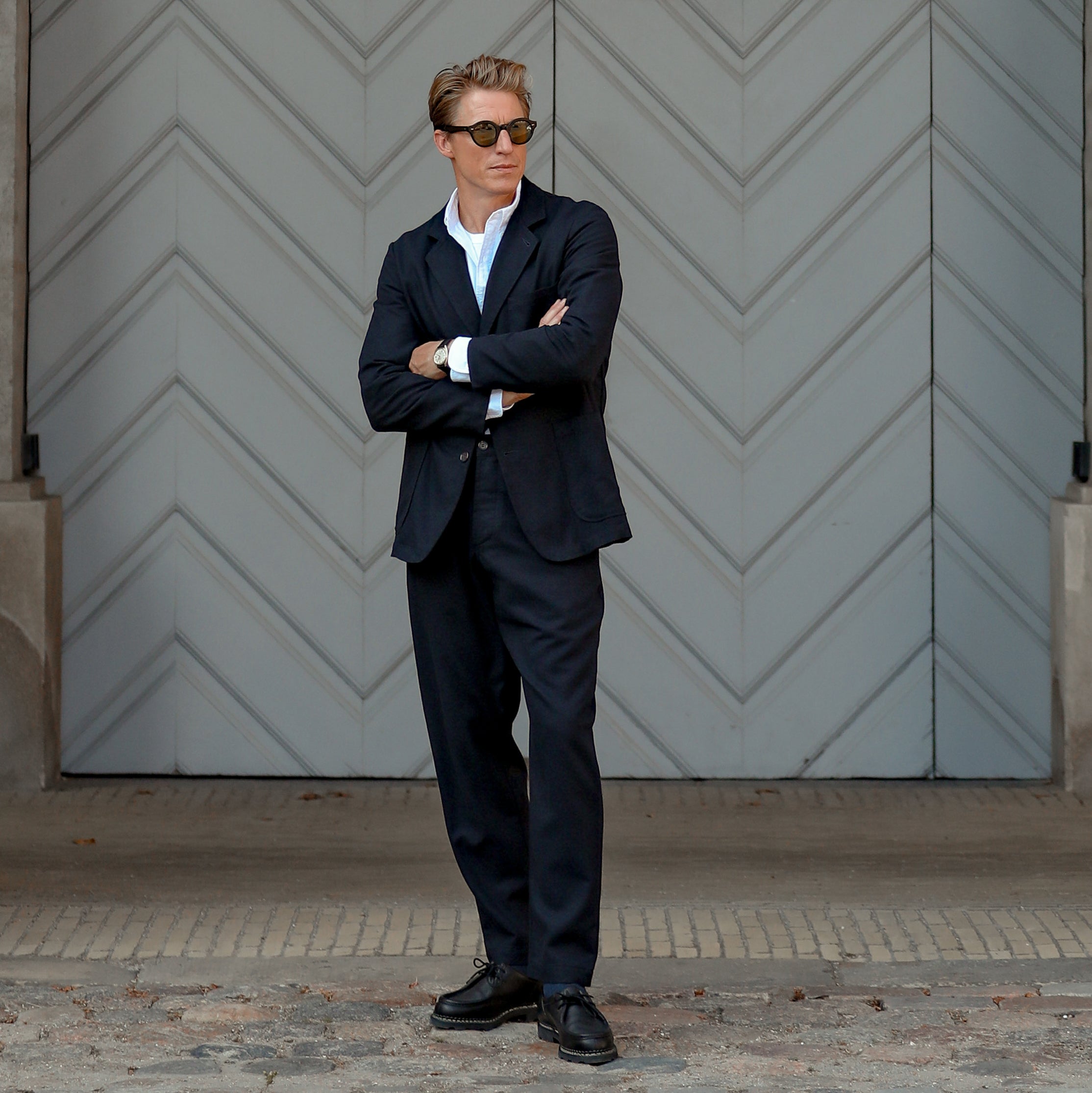 A person in a Navy Blue Upcycled Tweed 2 Button Jacket by Universal Works and navy blue sunglasses stands with arms crossed in front of a large, patterned, grey door.