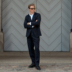 A person in a Navy Blue Upcycled Tweed 2 Button Jacket by Universal Works and navy blue sunglasses stands with arms crossed in front of a large, patterned, grey door.