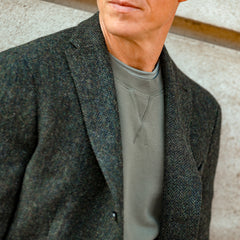 A person in a Khaki Green Cotton Loopback Sweatshirt by Sunspel and a tweed blazer stands against a light-colored wall.