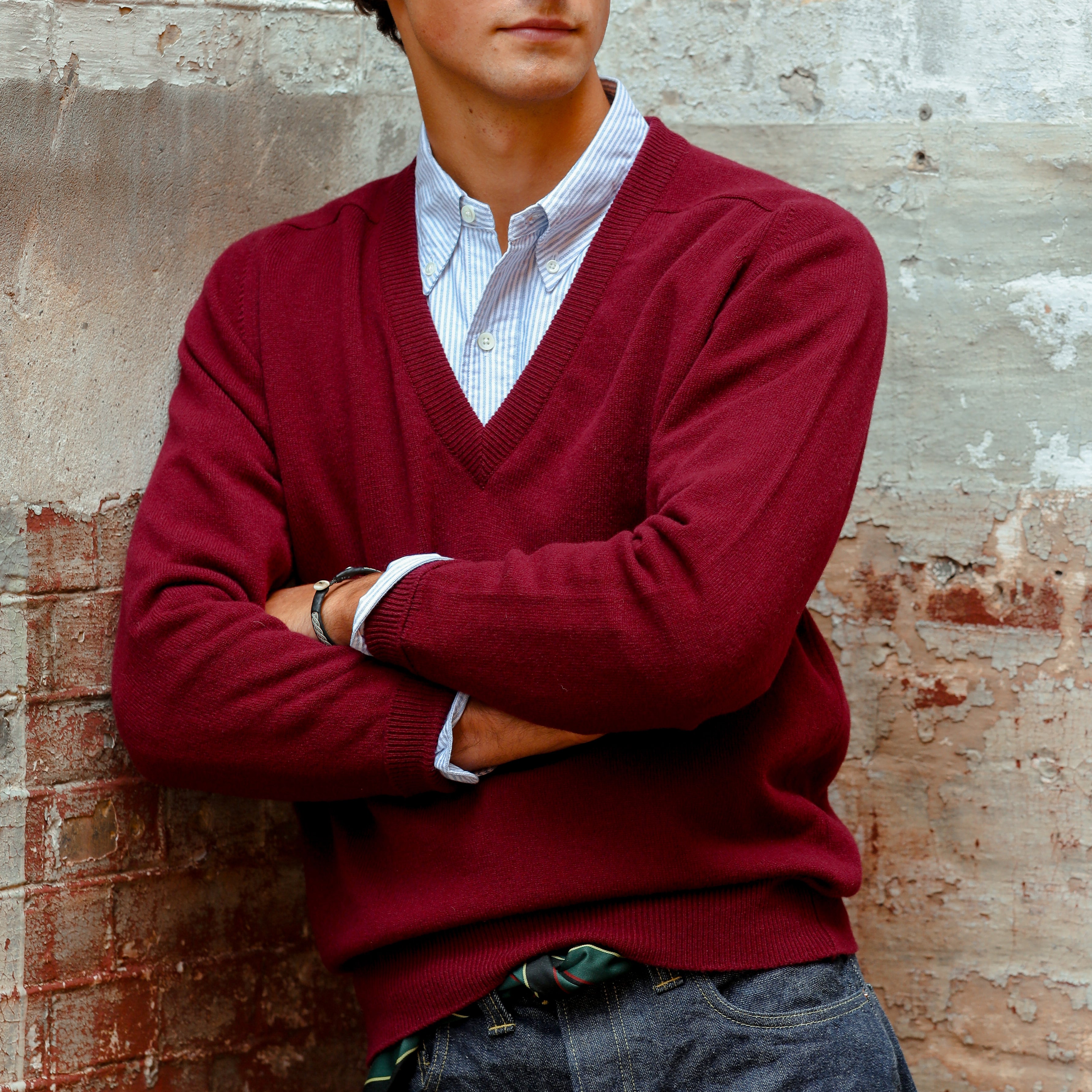 A person stands with arms crossed, wearing a William Lockie Bordeaux Deep V-Neck Lambswool Sweater over a light blue collared shirt, against a textured concrete and brick wall.