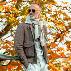 A man wearing sunglasses, a Valstar Taupe Suede Leather Shearling Flight Jacket, and a scarf stands in front of a tree with autumn leaves.