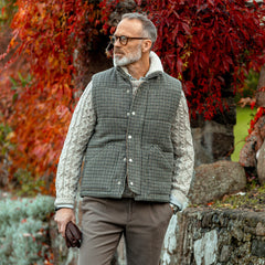 A man with a gray beard and glasses stands outdoors near a stone wall and red leaves, wearing a knit sweater beneath a Valstar Green Houndstooth Wool Tweed Padded Gilet, made in Italy.