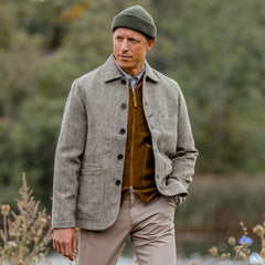 A person stands outdoors wearing a green beanie, grey button-up coat, brown zip-up sweater, and beige pants with hands in pockets. The outfit is accentuated by the Mustard Yellow Wool Fleece Zip Waistcoat from Universal Works. The background features greenery with some flowers in the foreground.