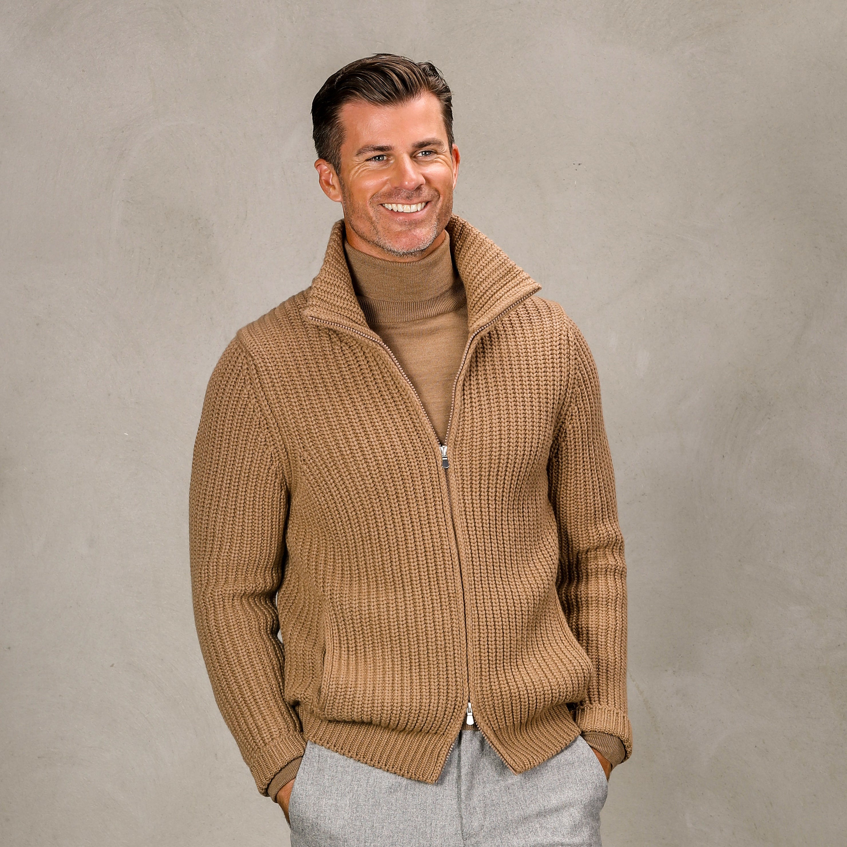 A man is wearing a Gran Sasso Camel Wool Cashmere Heavy Knit Cardigan over a tan turtleneck. He has light brown hair and is smiling, standing against a plain, textured gray background.