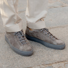 Close-up of a person's feet wearing CQP Granit Grey Suede Leather Racquet Sr sneakers and beige trousers on a paved surface.